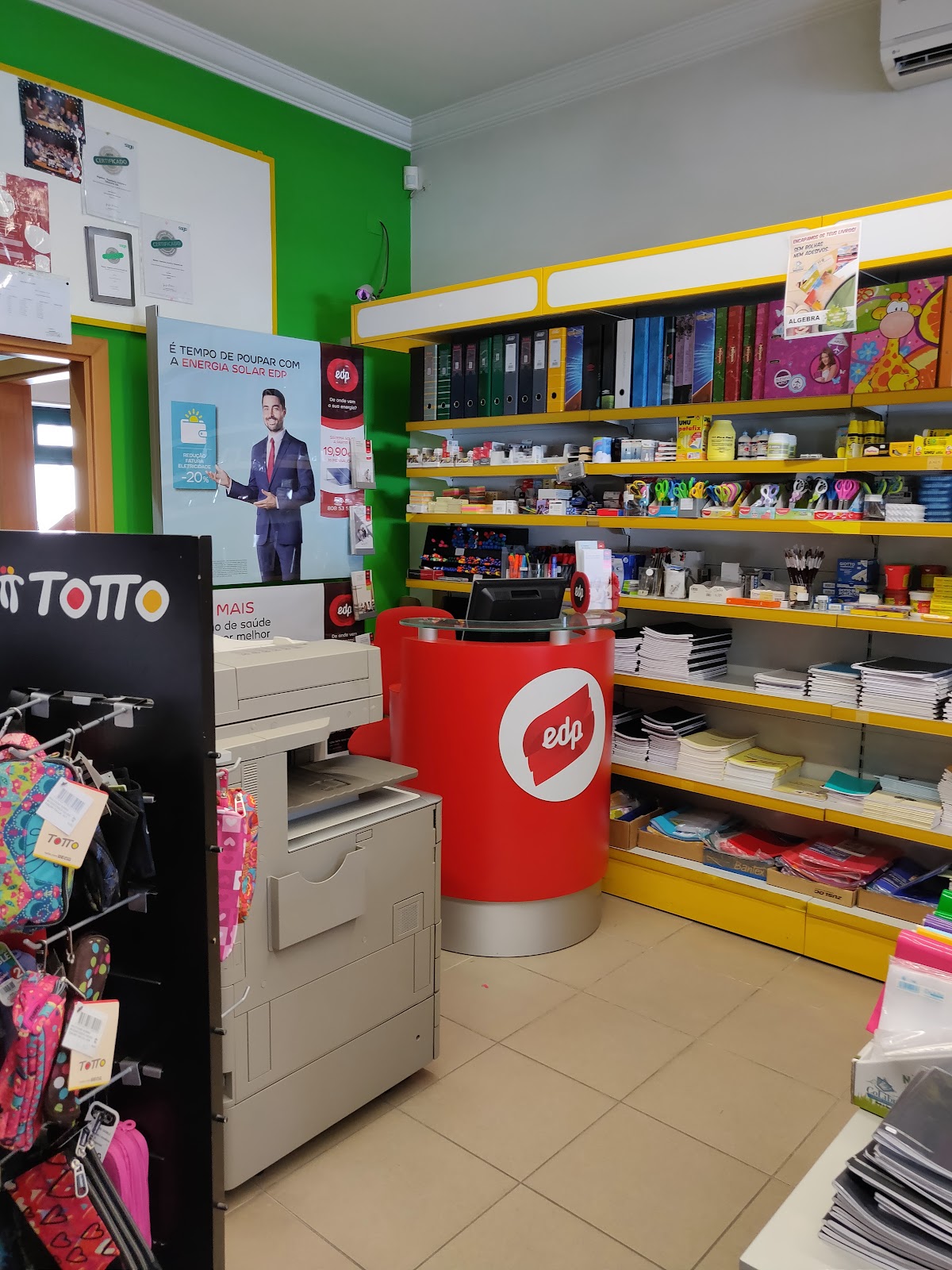 Interior da papelaria: estantes amarelas com cadernos, material escolar e mochilas